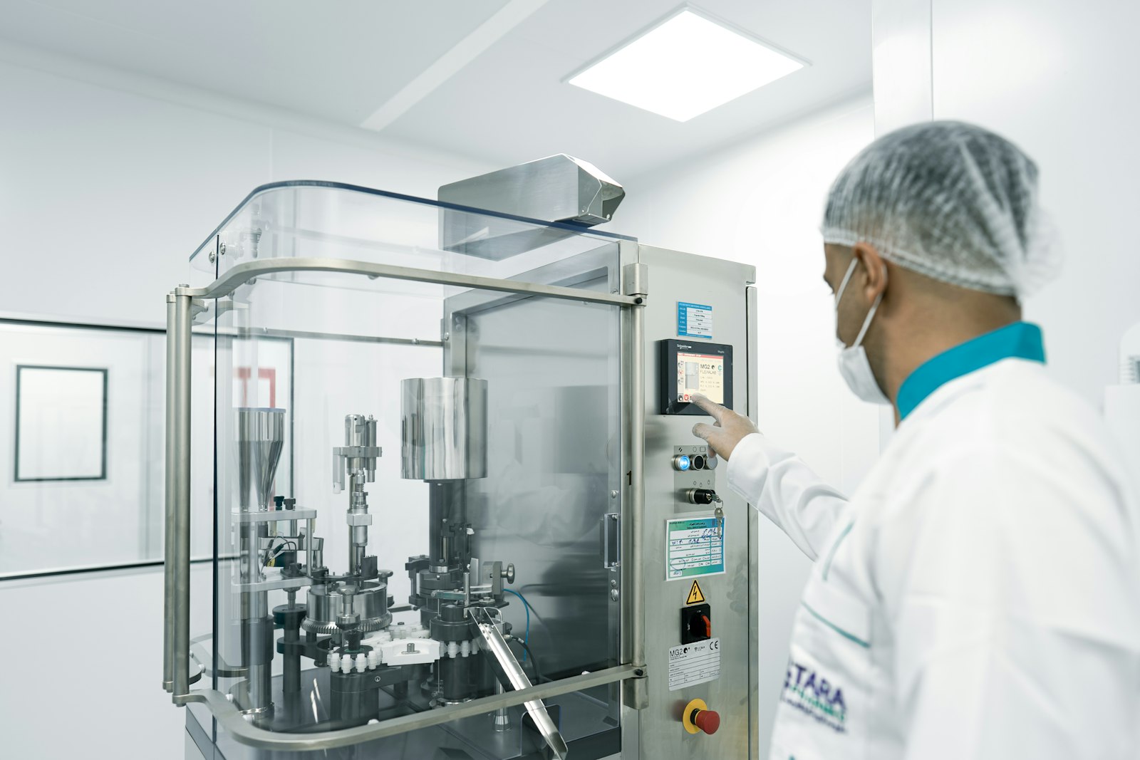 Operator in a hairnet and gown beside pharmaceutical production equipment